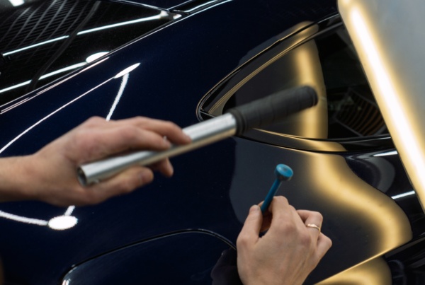 A technician performs paintless dent repair on a dark blue car door, using a metal rod and light-reflecting board from behind while gently tapping the panel with a blue-handled tool from the front to gauge dent depth.