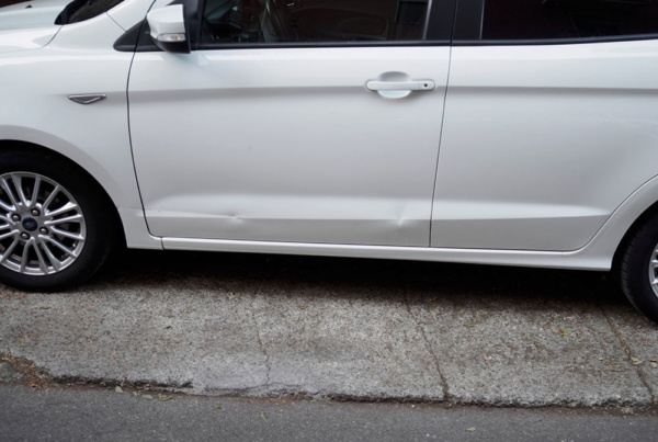 White Ford hatchback parked on a sloped curb, showing multiple dents along the lower part of the driver-side front door.